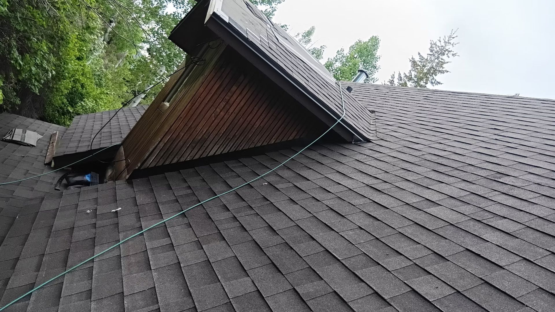A dark shingle roof with a damaged section and a chimney-like structure against green trees.