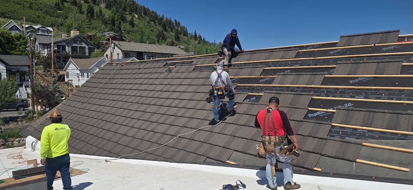 Roofers working on a roof, installing shingles on a sunny day. Mountain and houses in the background.