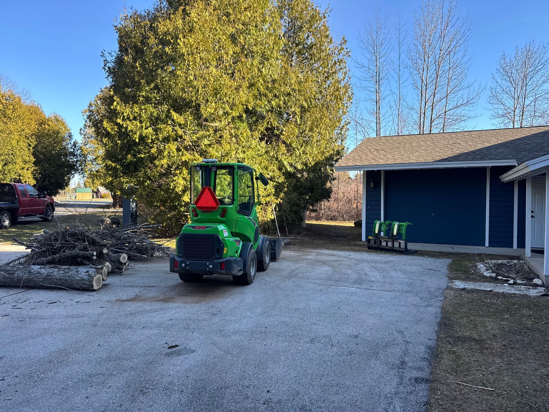 Green tractor on gravel driveway near wood pile and blue building.