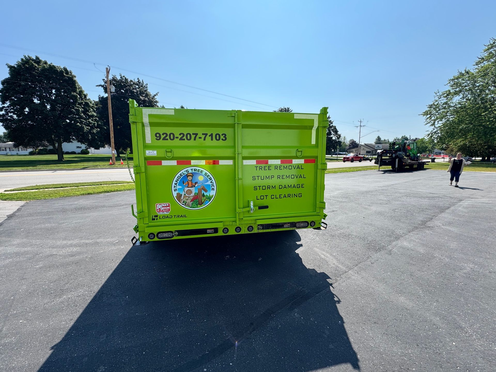 Green dumpster on asphalt with contact info, two people, sunny day.