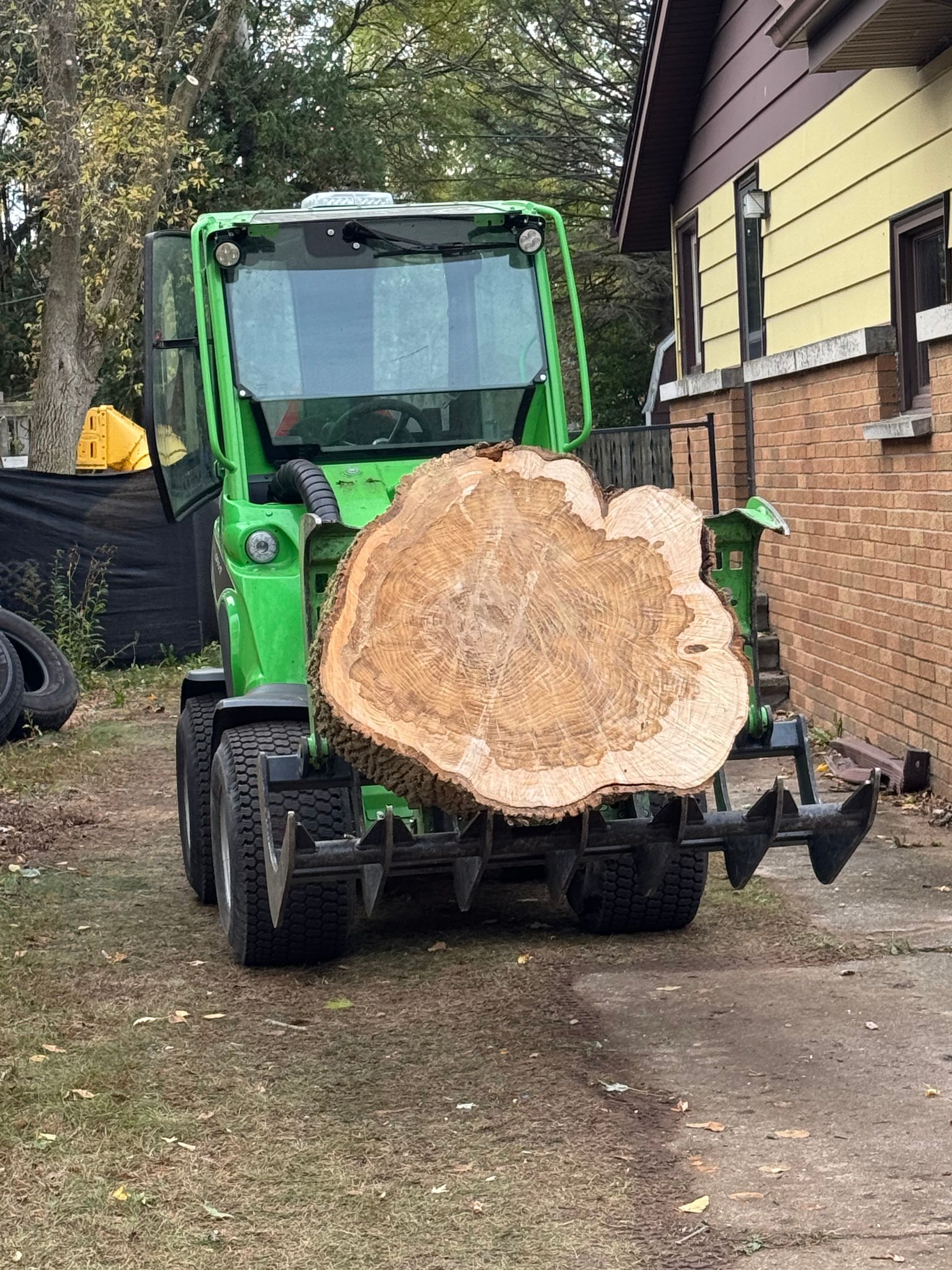 Green compact loader holding a large tree slice outdoors near a brick building.
