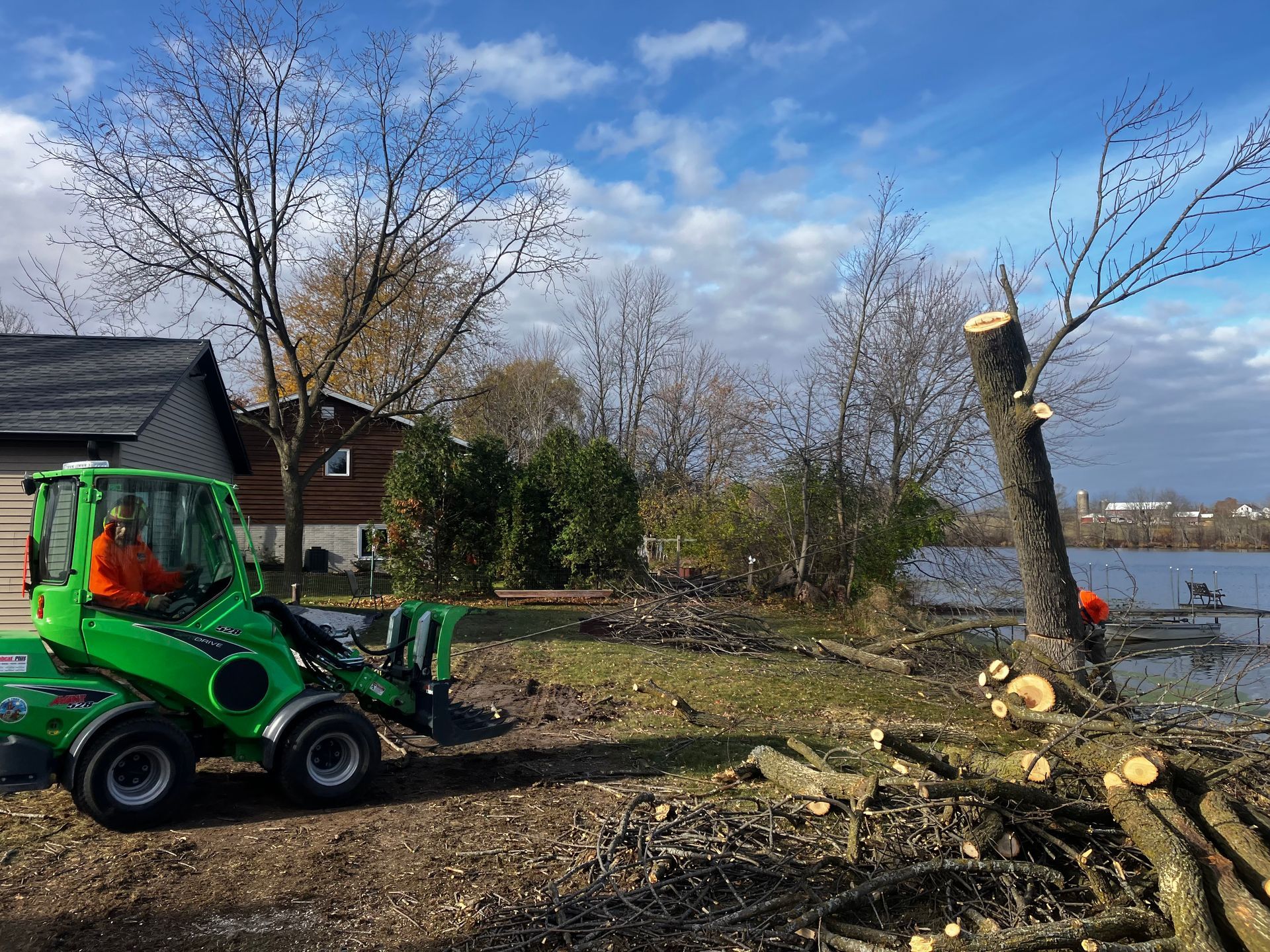A person operates a green tractor clearing branches near a lake on a sunny day.