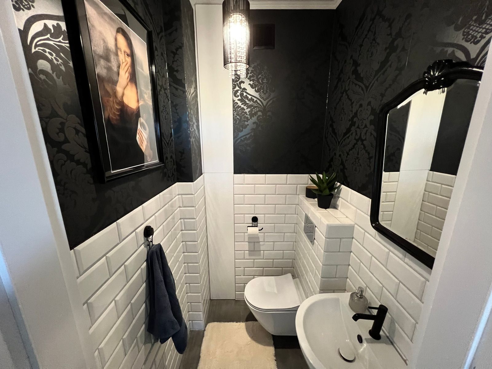 A small, modern bathroom featuring black damask wallpaper, white subway tiles, a wall-mounted toilet, and a sink.