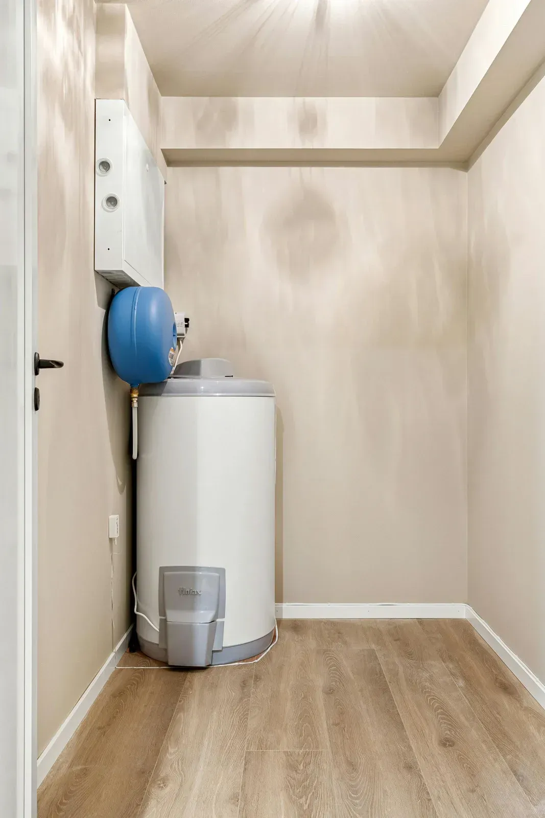 A white hot water tank and blue expansion vessel installed in a small, empty utility room with light wood-look flooring.