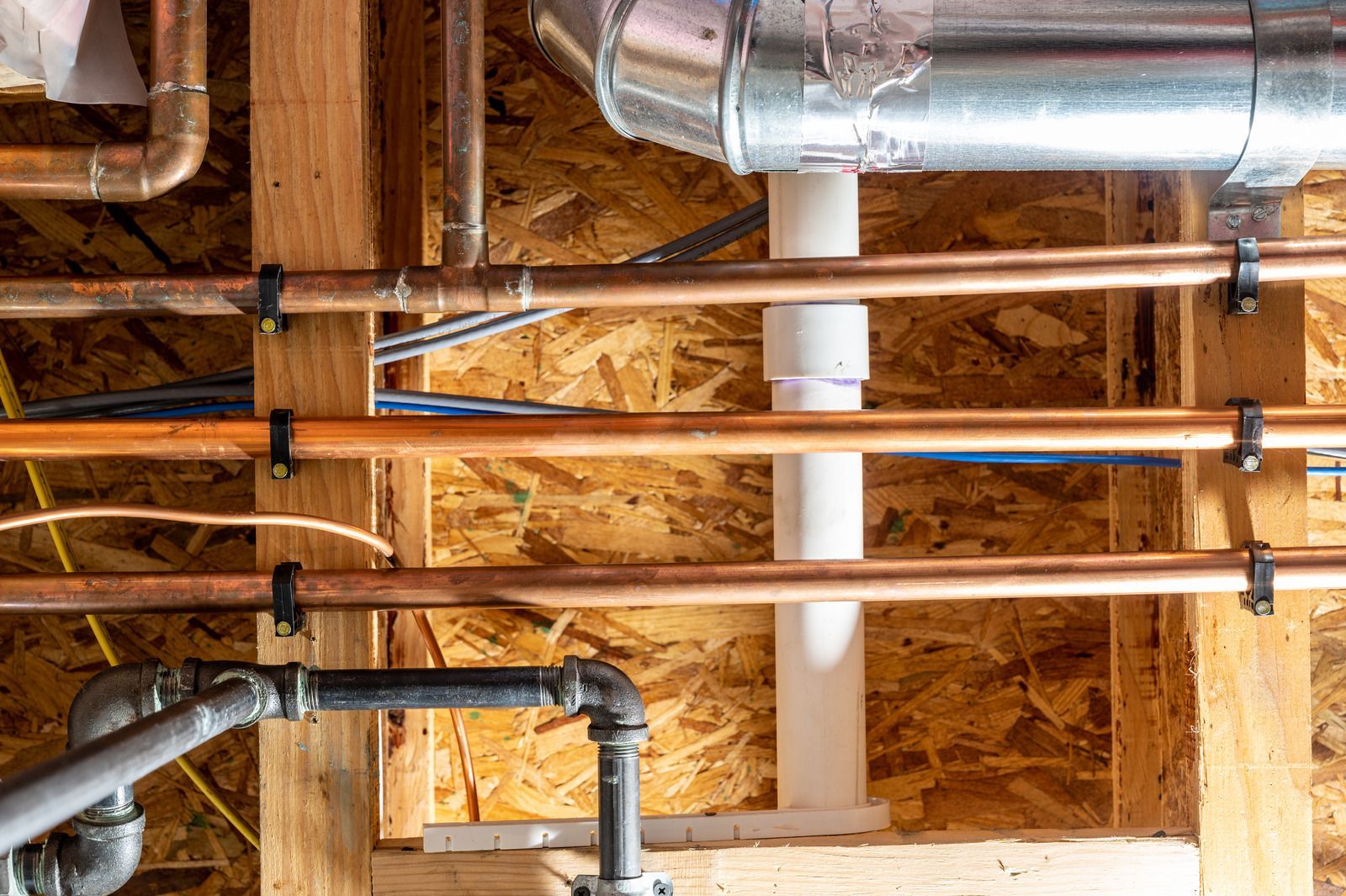 Copper pipes, a black metal pipe, and a white plastic pipe installed under wooden ceiling joists and ductwork.