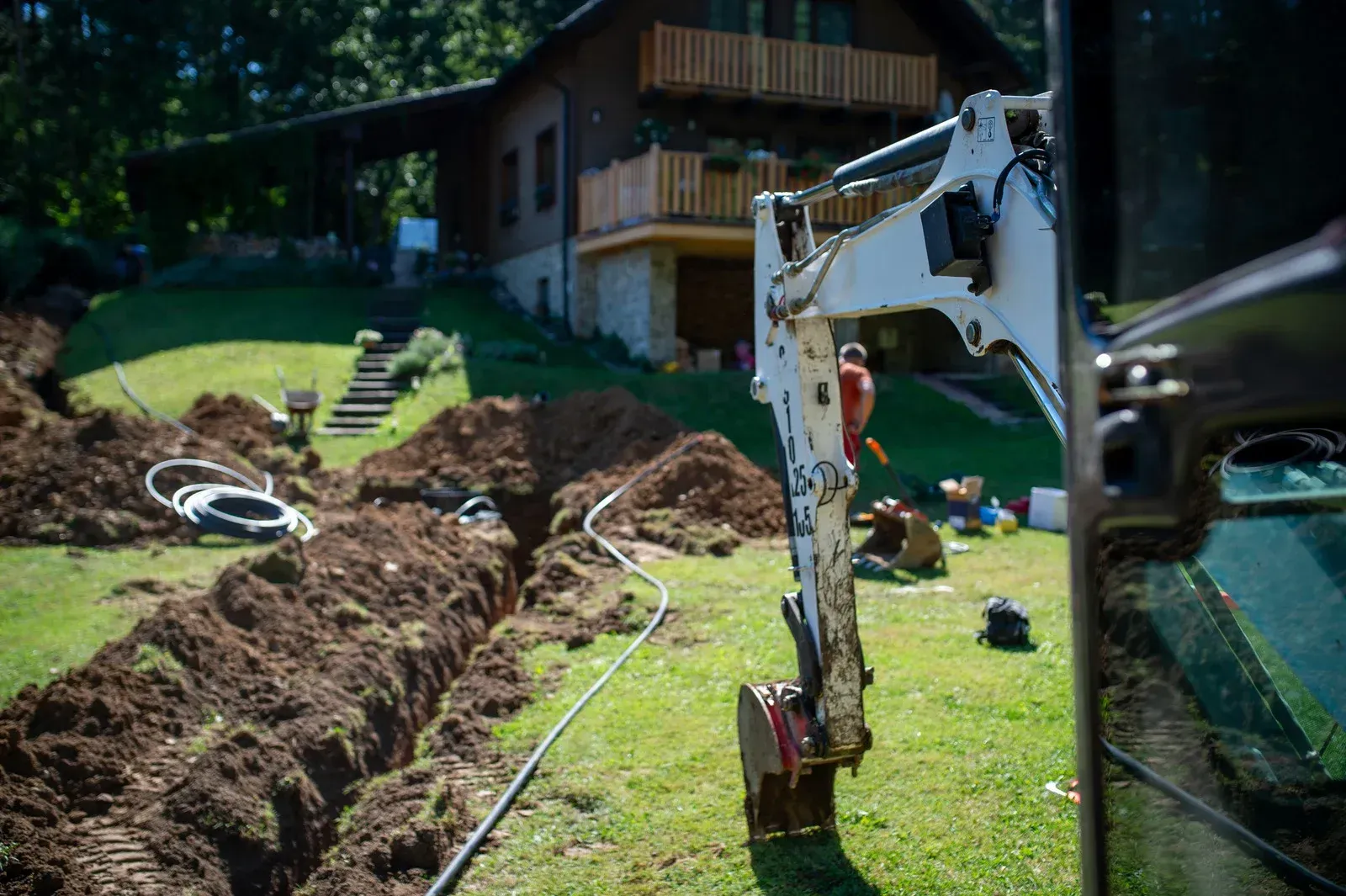 An excavator arm stands in a green yard next to a deep trench, with a house in the background.