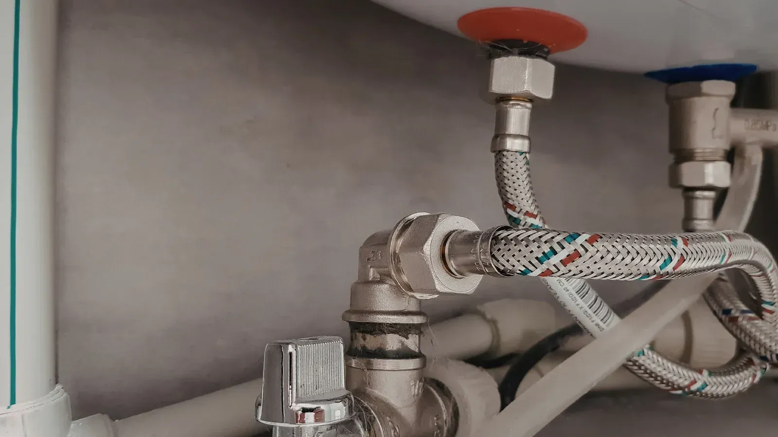 Close-up of plumbing connections under a sink, showing braided metal hoses, a chrome shut-off valve, and pipes.