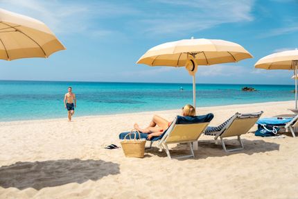 Woman relaxes on beach chair under umbrella as man walks on the shore; turquoise water and blue sky.