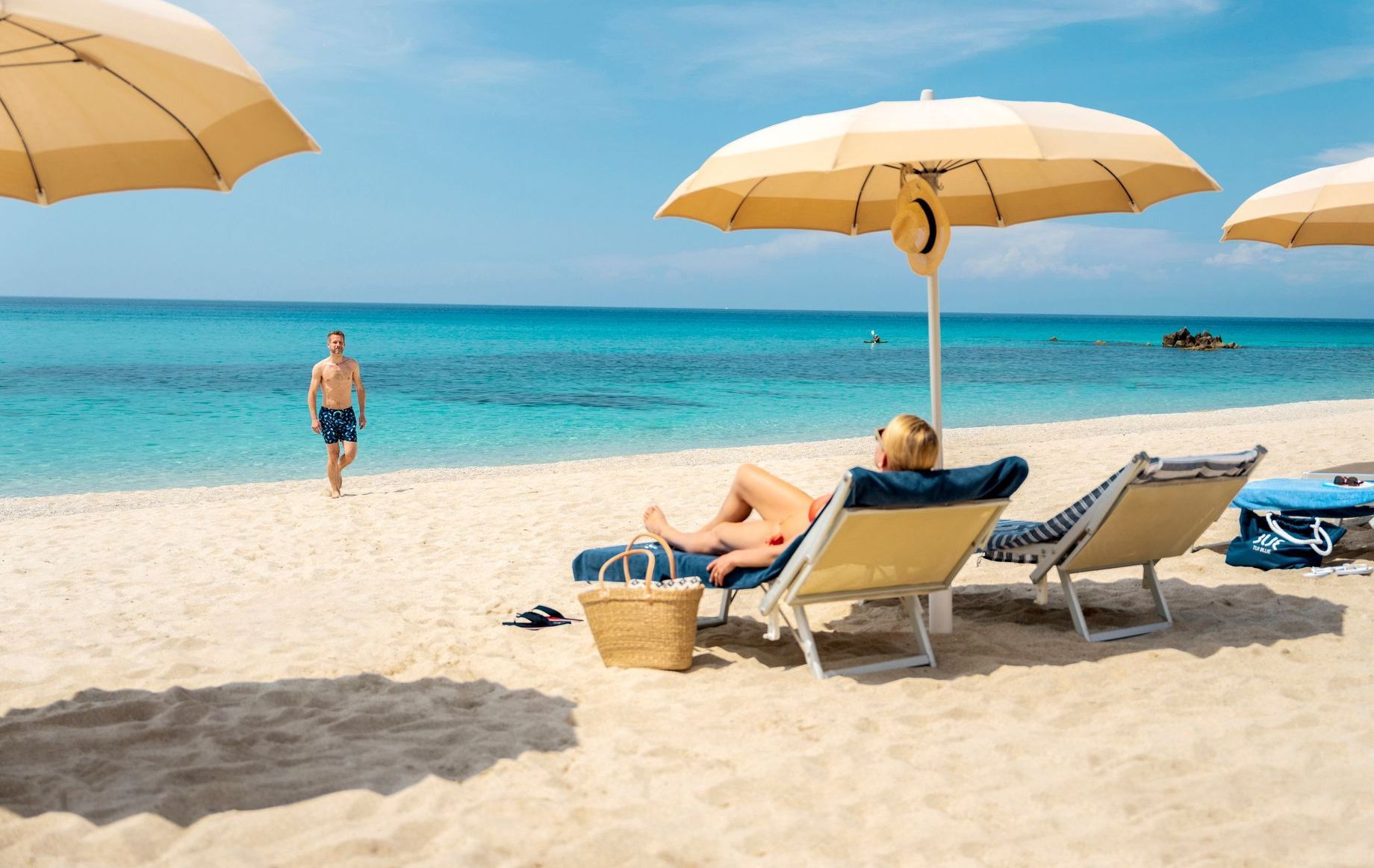 Person relaxing on a beach chair under an umbrella, with a man walking nearby, blue ocean, white sand.