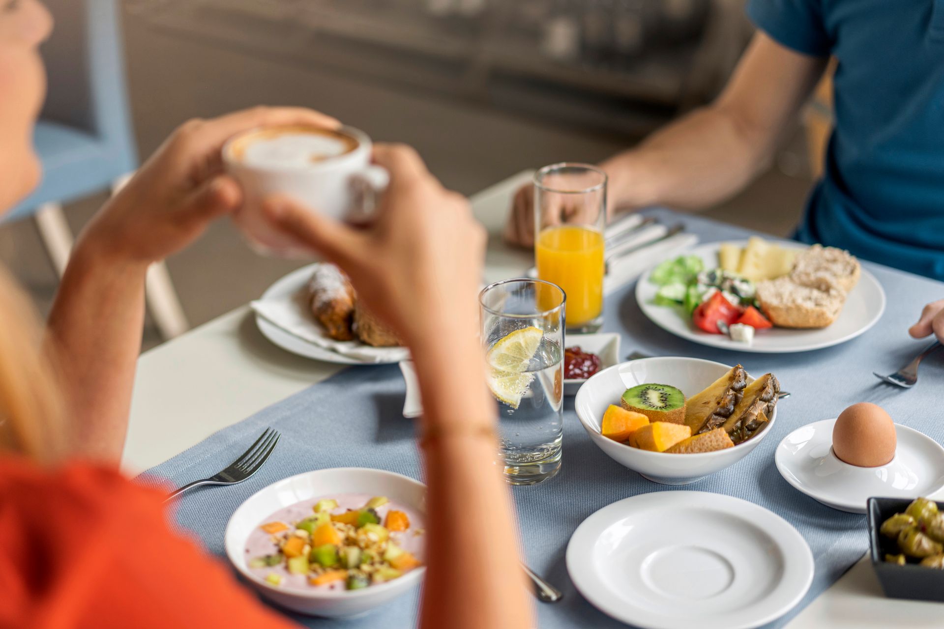 People eating breakfast at a table: coffee, juice, fruit, bread, egg, and olives.