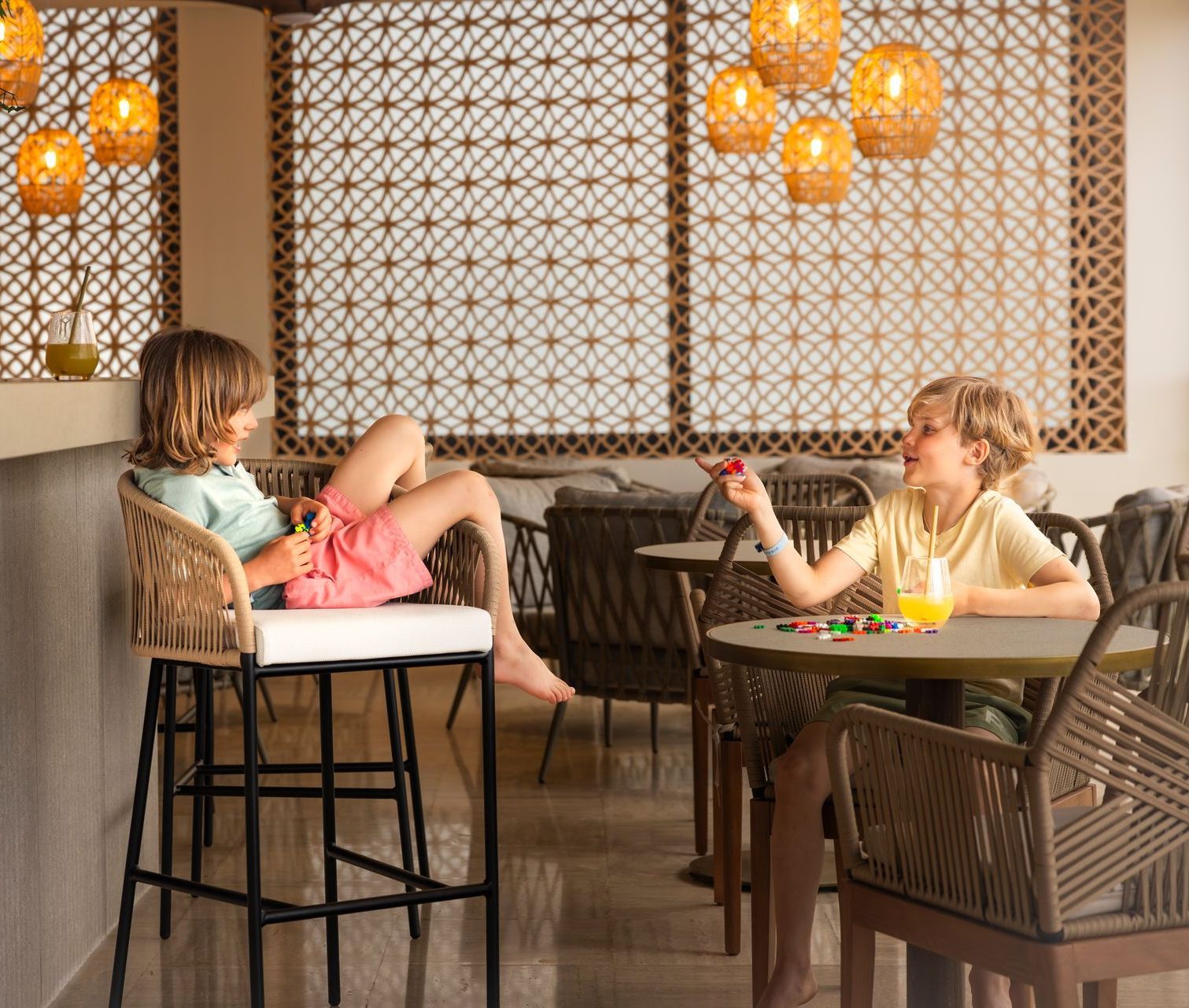 Two children play at a cafe. One sits on a stool, the other at a table. Light fixtures and patterned screen.