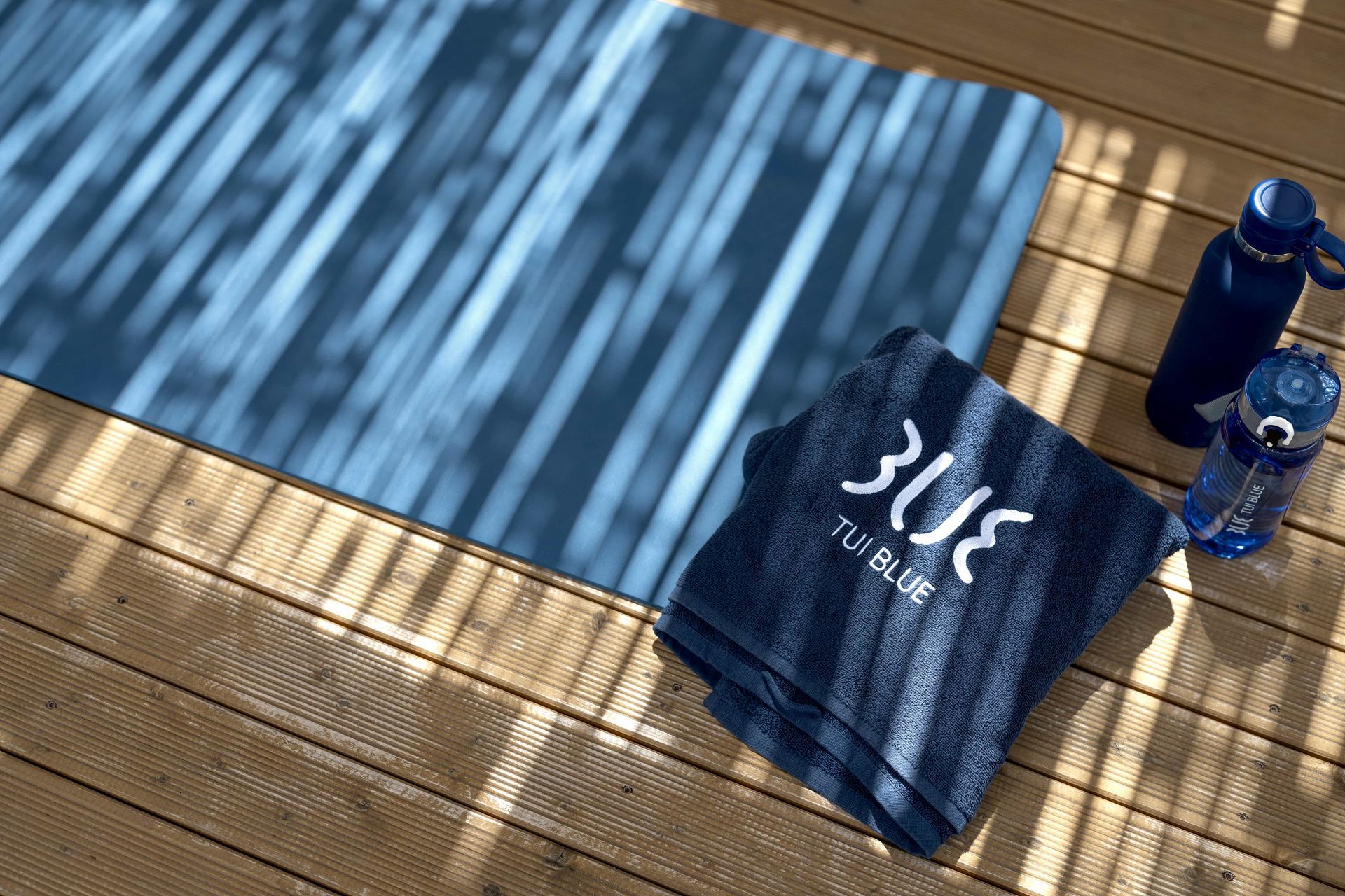 Blue yoga mat, towel, and water bottles on a wooden deck, lit by shadows.