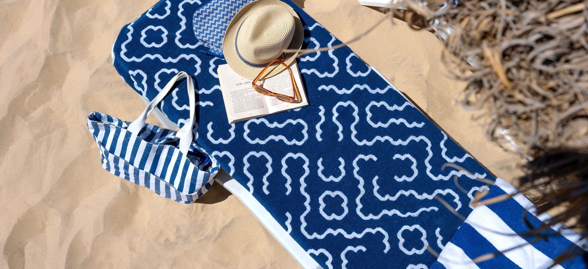 Beach scene: a blue patterned towel, hat, bag, and reading material on sand.