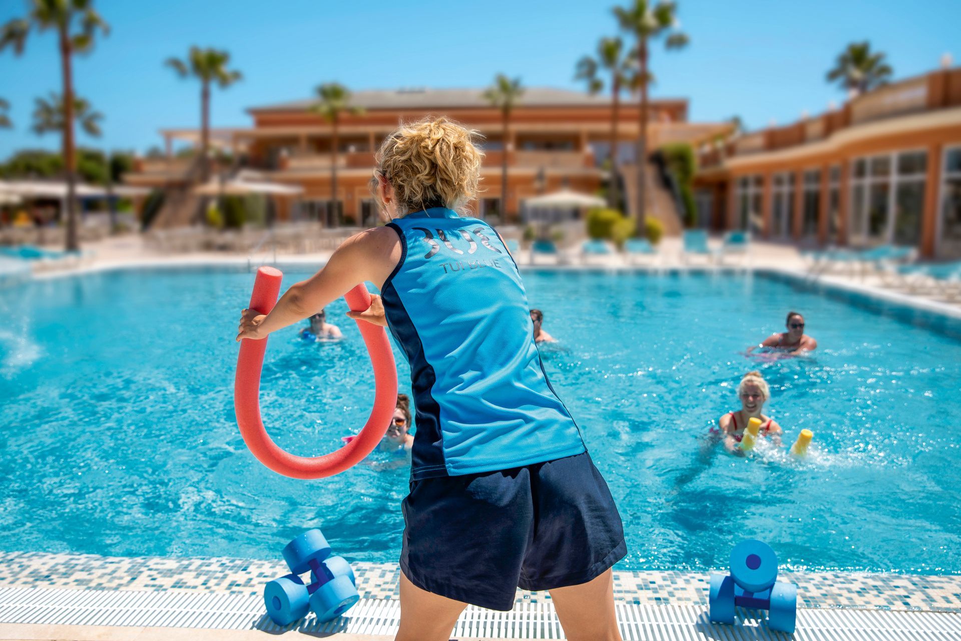 Woman leading water aerobics class in a pool, holding a red pool noodle, with other people in the water.