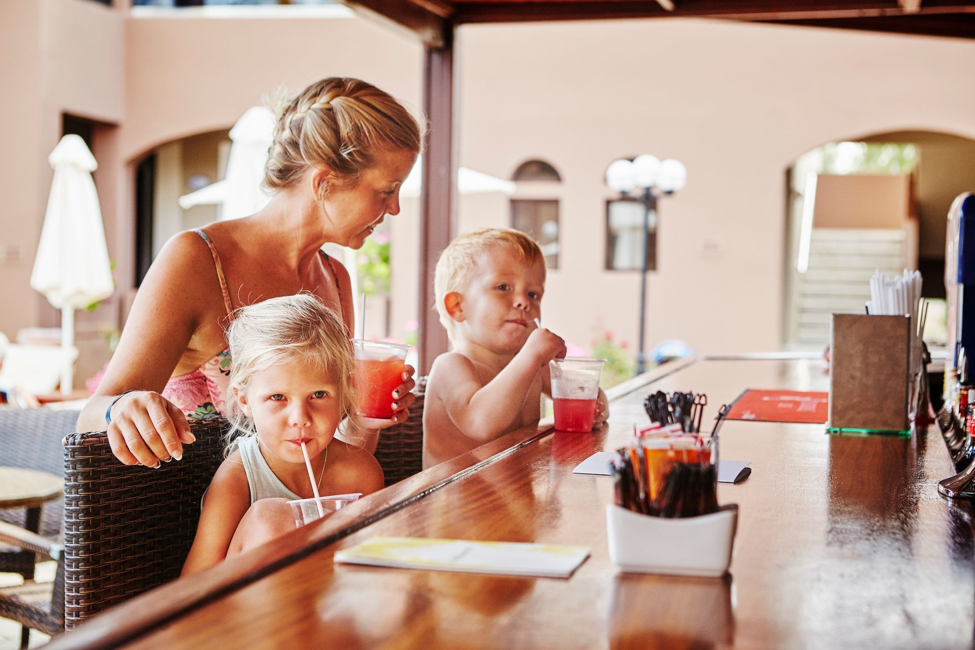 Woman and two children at a bar, drinking red beverages. Blond hair, outdoor setting.