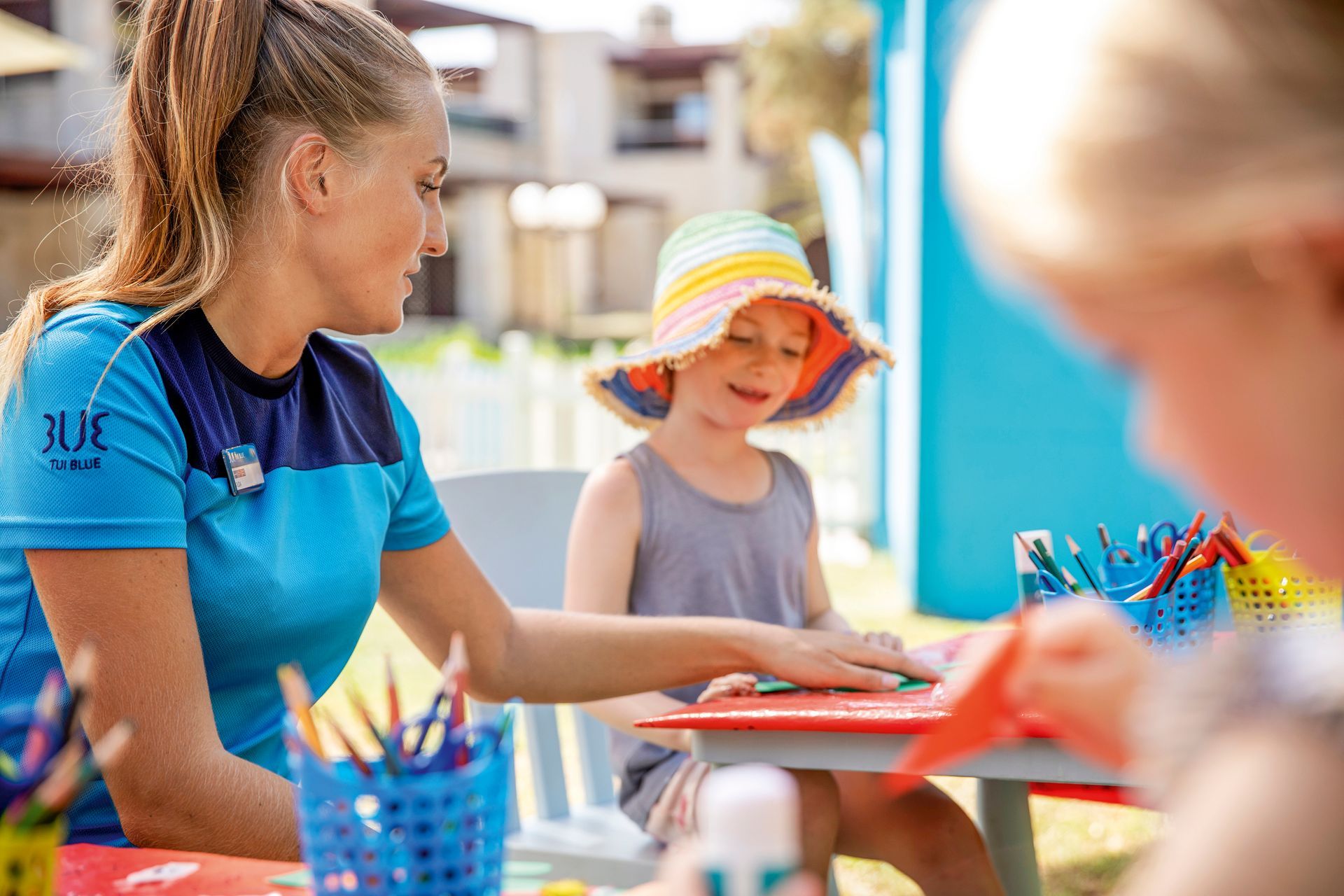Woman assists a child at a craft table with colored pencils. Sunny outdoor setting.