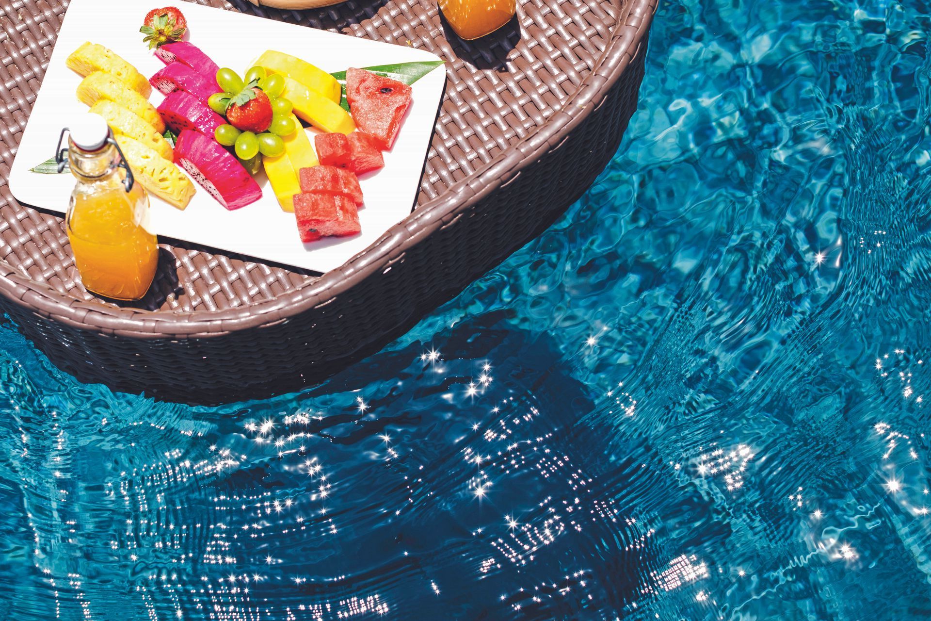 Floating tray with fruit and juice in a pool of blue water.