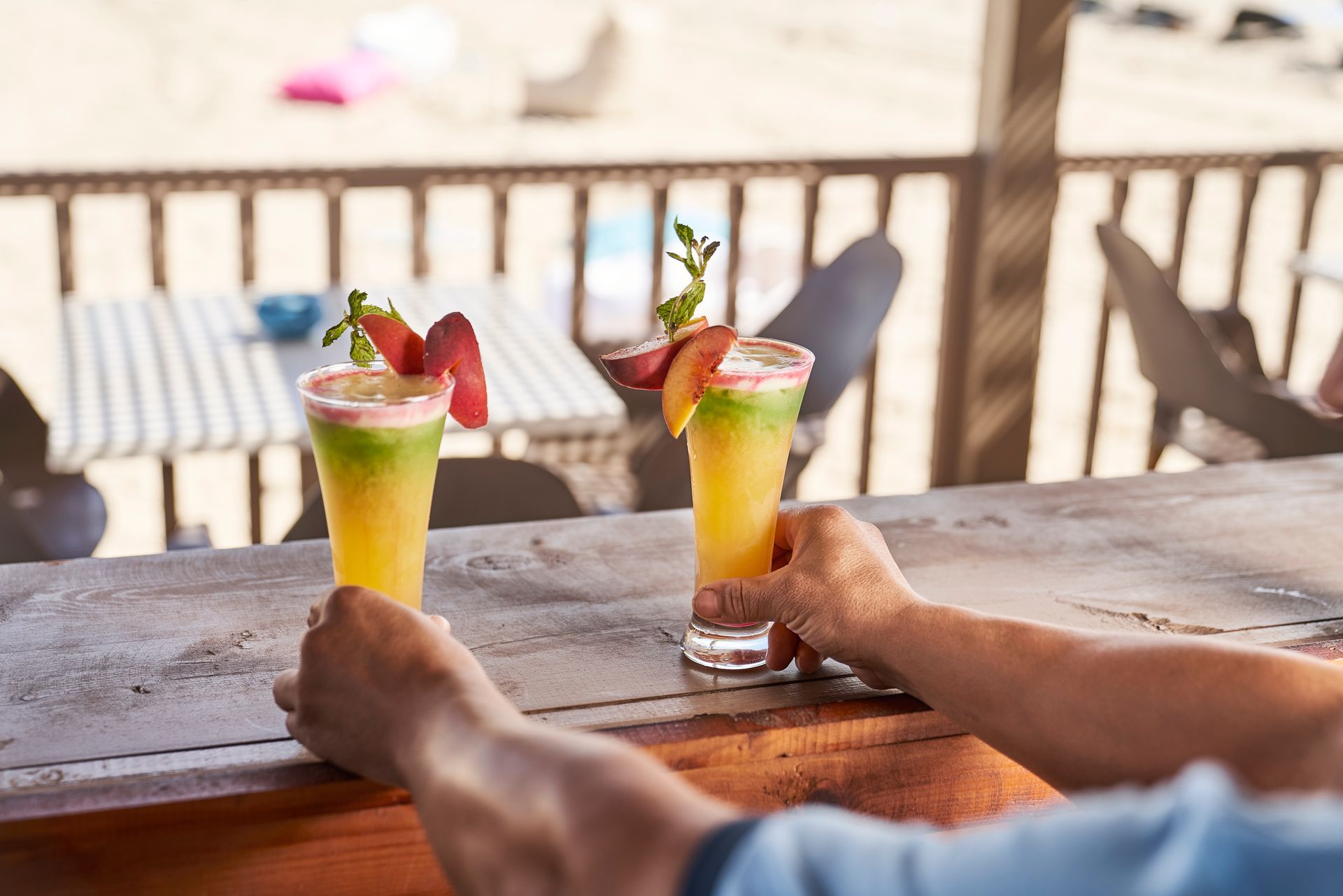 Two cocktails garnished with fruit, held by hands on a bar with beach scenery in the background.