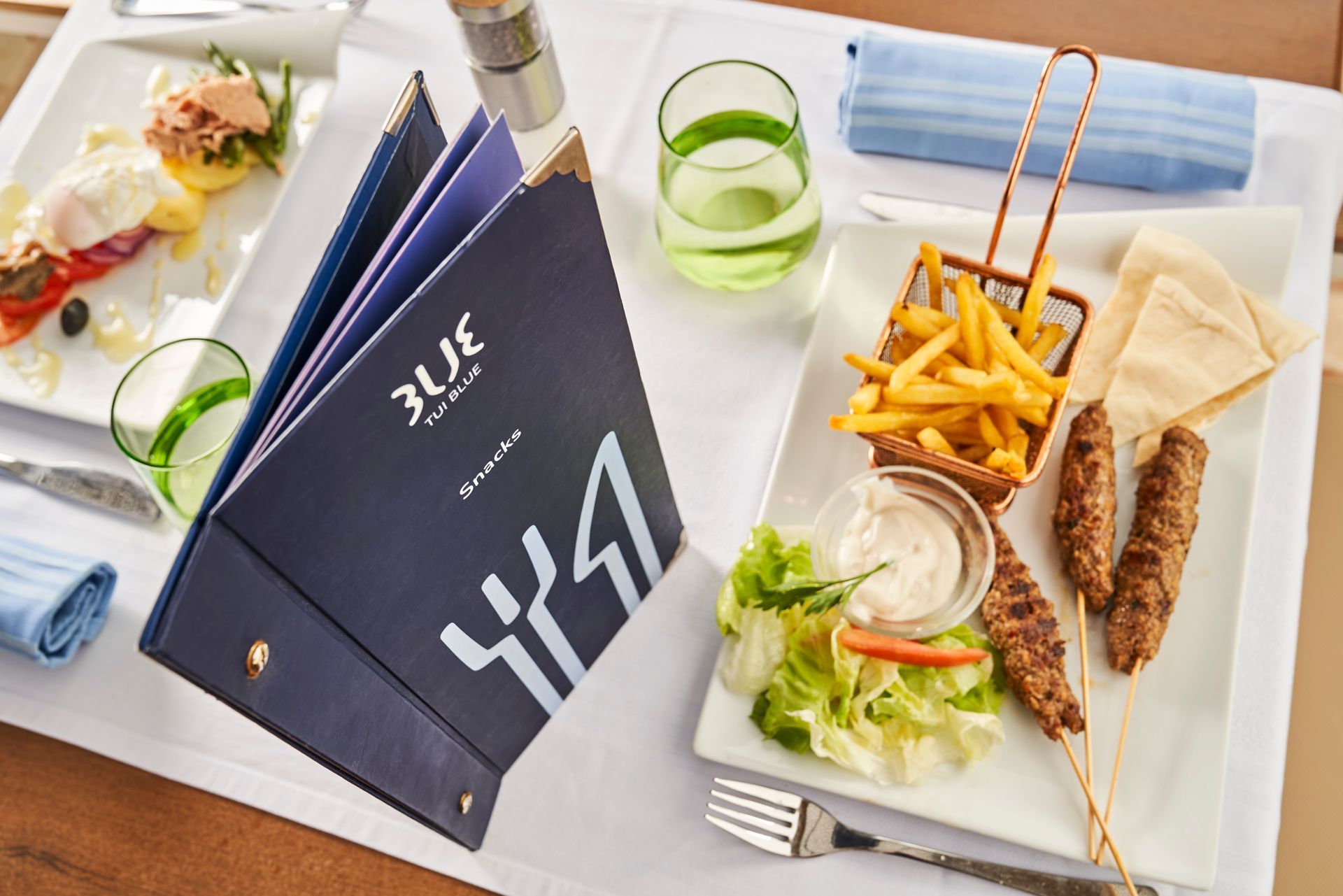 Blue menu with food: fries, kebabs, and salad on a white table.