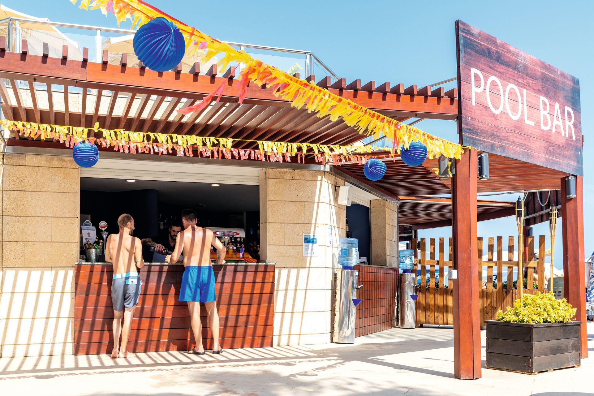 Pool bar with two men in swim trunks, sunlight, wood structure, festive decorations.