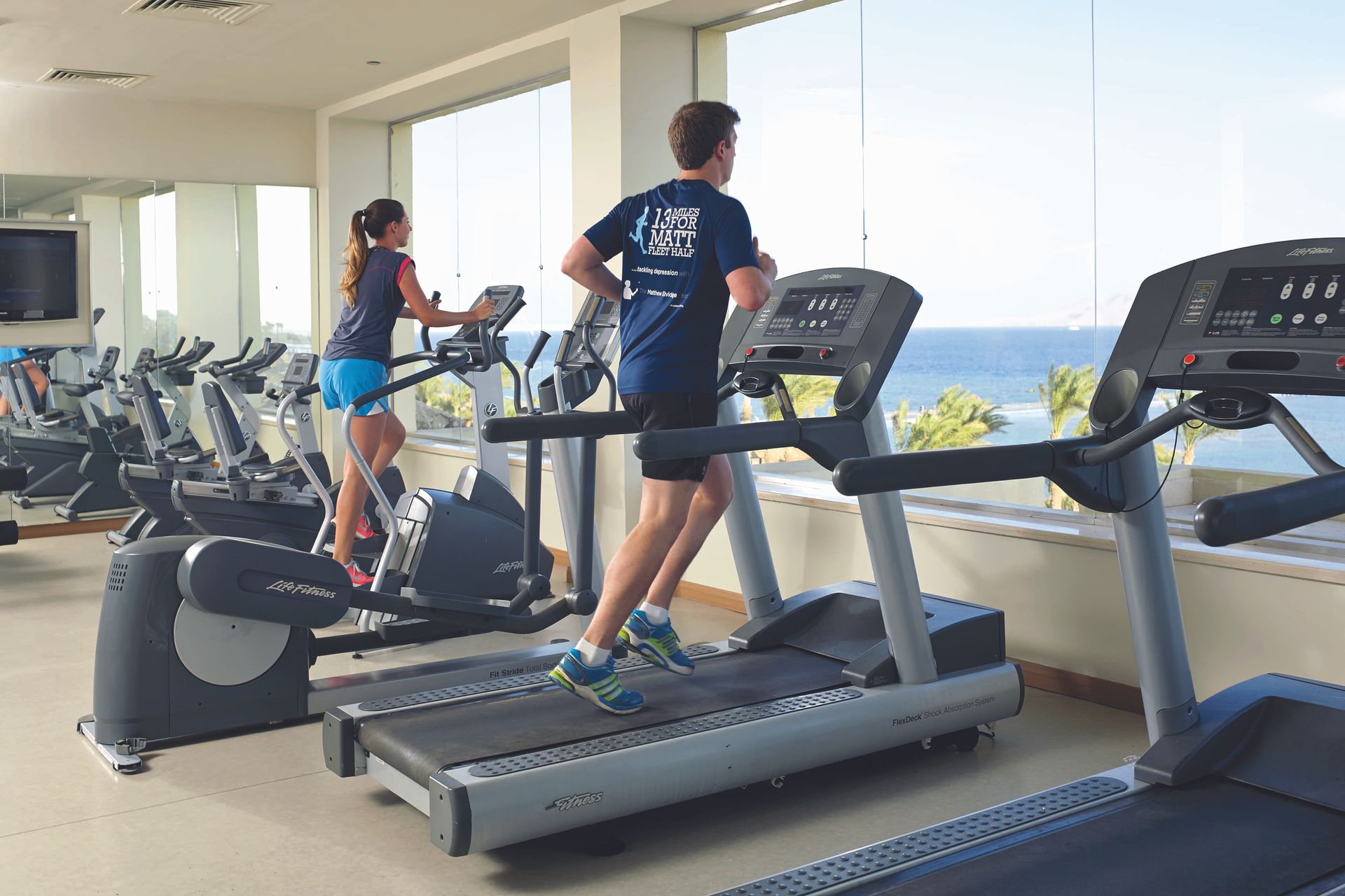 Man runs on treadmill, woman uses elliptical in gym overlooking ocean.