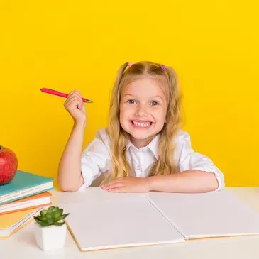 Smiling young girl holding a pencil, sitting at a desk with a red apple.