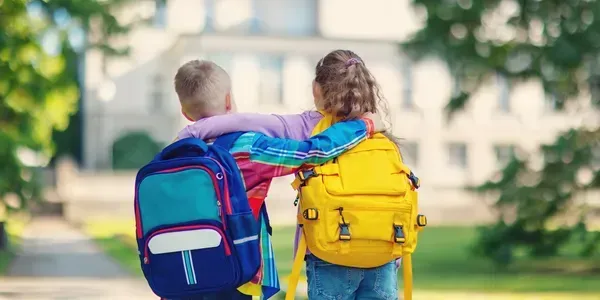 Two children walking side by side with colorful backpacks, arms around each other.