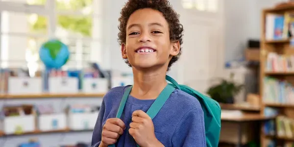 Young student smiling with a blue backpack in a classroom setting.