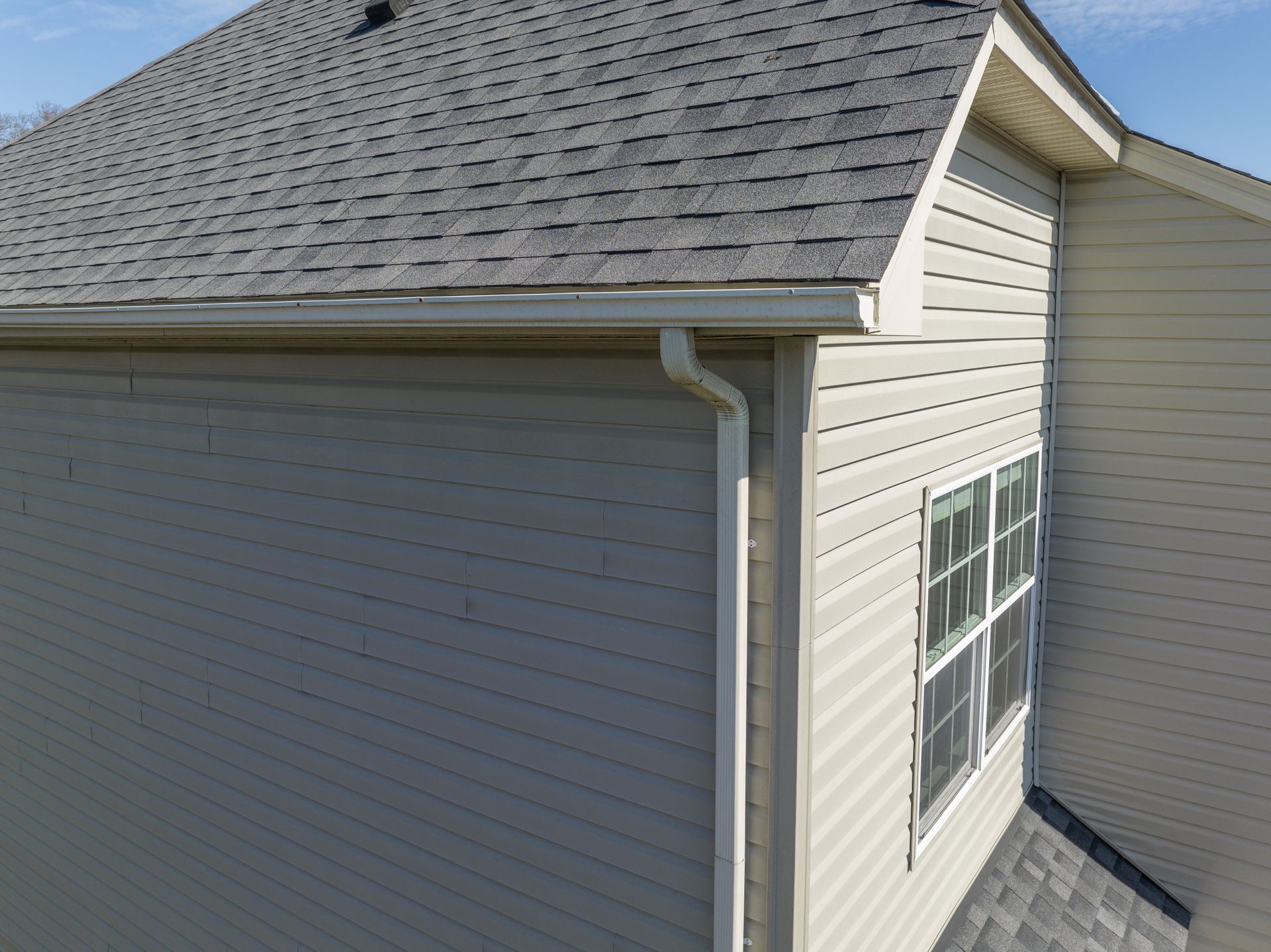 Exterior corner of a beige house with gray shingle roof and white trim, showing a window and gutter