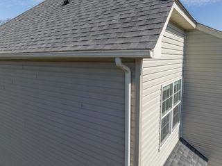 Corner of a gray house with vinyl siding, shingled roof, gutters, and a white-trimmed window.