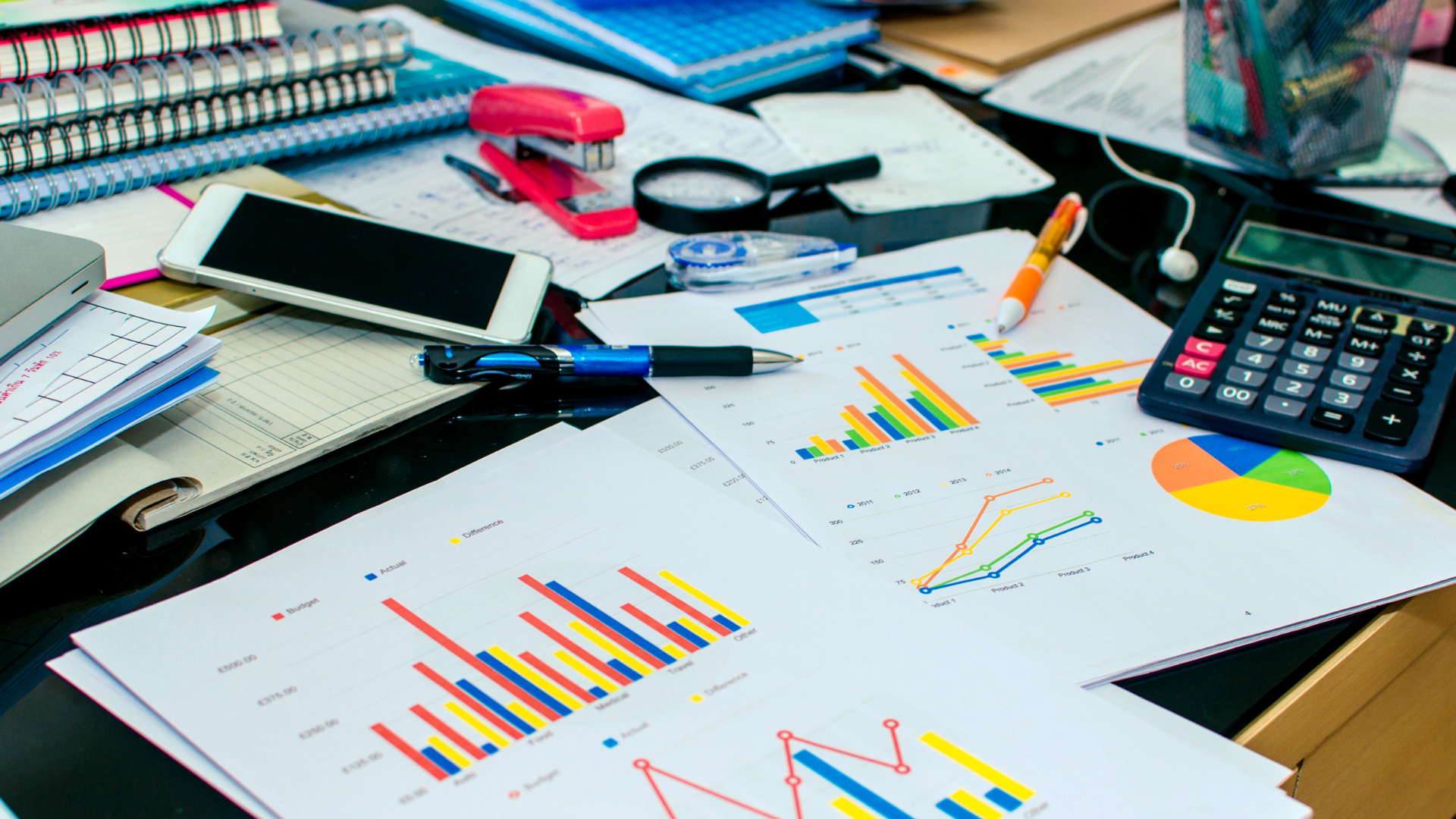 A messy desk with papers , a calculator , a cell phone , and a magnifying glass.