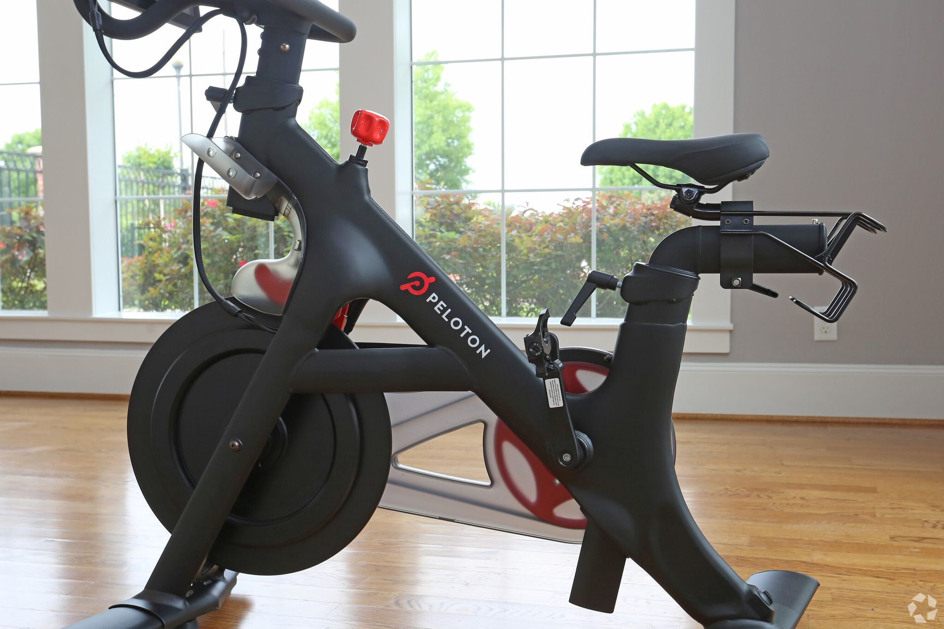 A peloton bike is sitting on a wooden floor in front of a window.