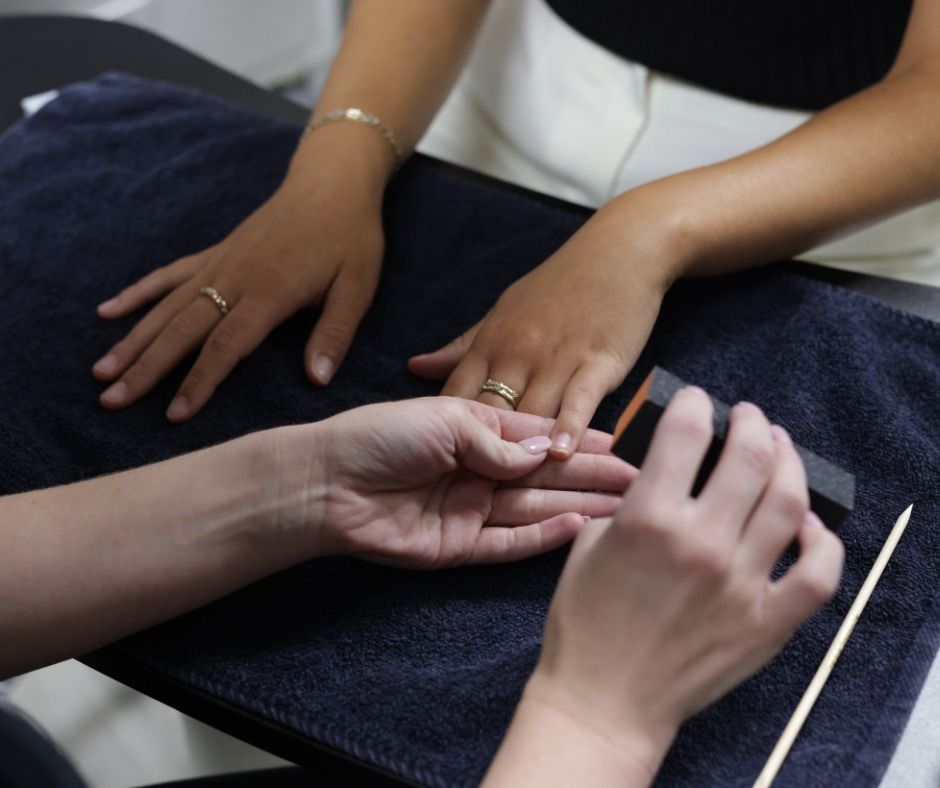 A manicurist uses a sanding block to file the fingernails of a client resting their hands on a dark blue towel.