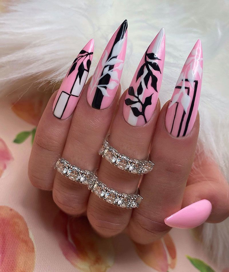 Long pink stiletto nails with black and white floral designs. The hand also wears diamond rings.