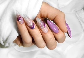 A hand with almond-shaped nails featuring purple gradient, marble-style art, and solid lilac polish against white fabric.