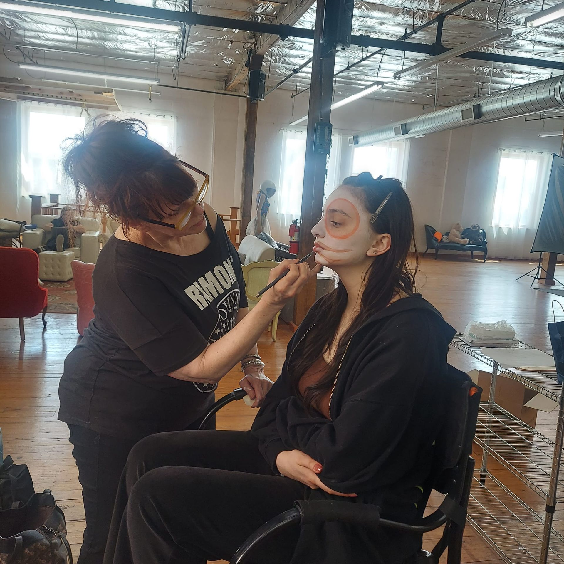 A makeup artist applying white face paint to a person sitting in a salon chair in a large, bright studio.