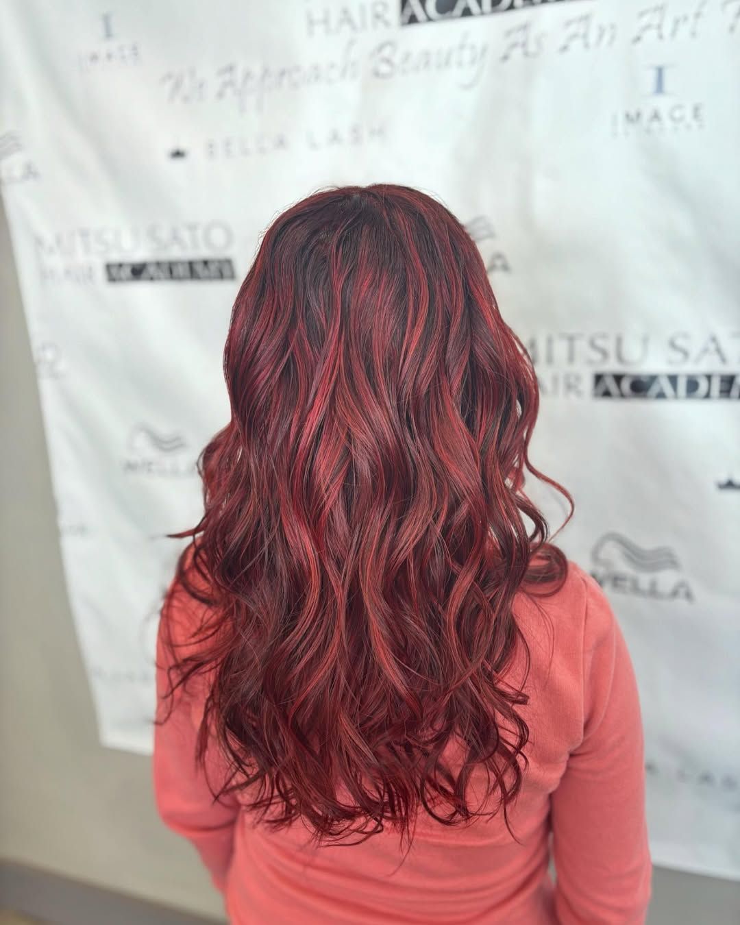 Woman with long, wavy, dark red hair. She wears a peach-colored top, standing in front of a white wall with writing.