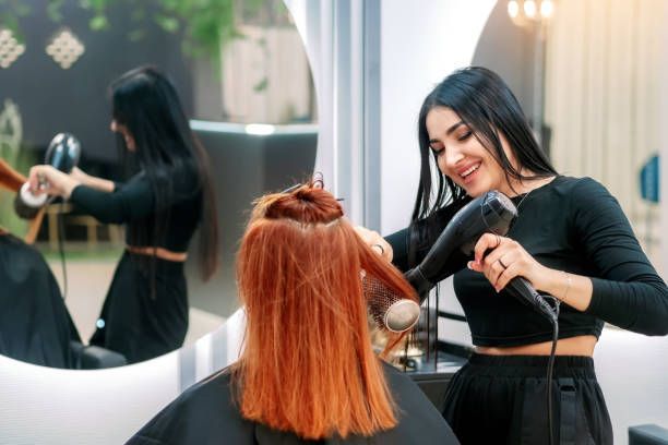 Hairdresser blow-drying a client's red hair in a salon; mirror reflects the process.