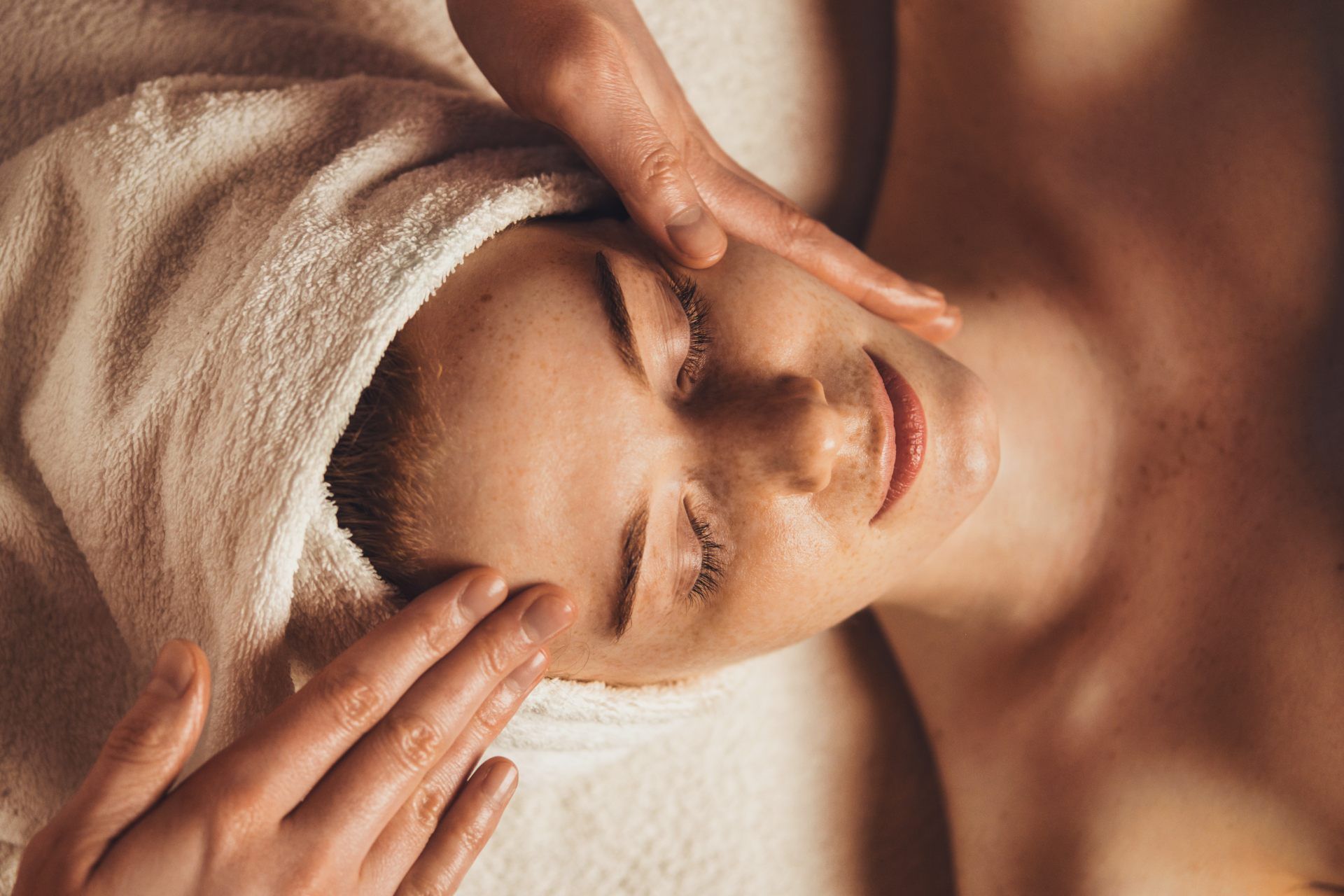 A person receives a gentle face massage while lying down with a white towel wrapped around their head.