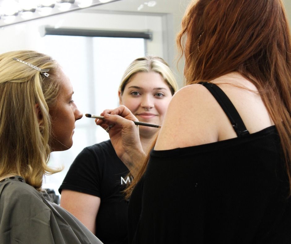 A makeup artist applies makeup to a person’s face while a woman watches from behind them in a mirror reflection.