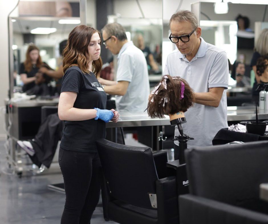 Stylist demonstrates hair coloring on a mannequin with student watching in a modern salon.