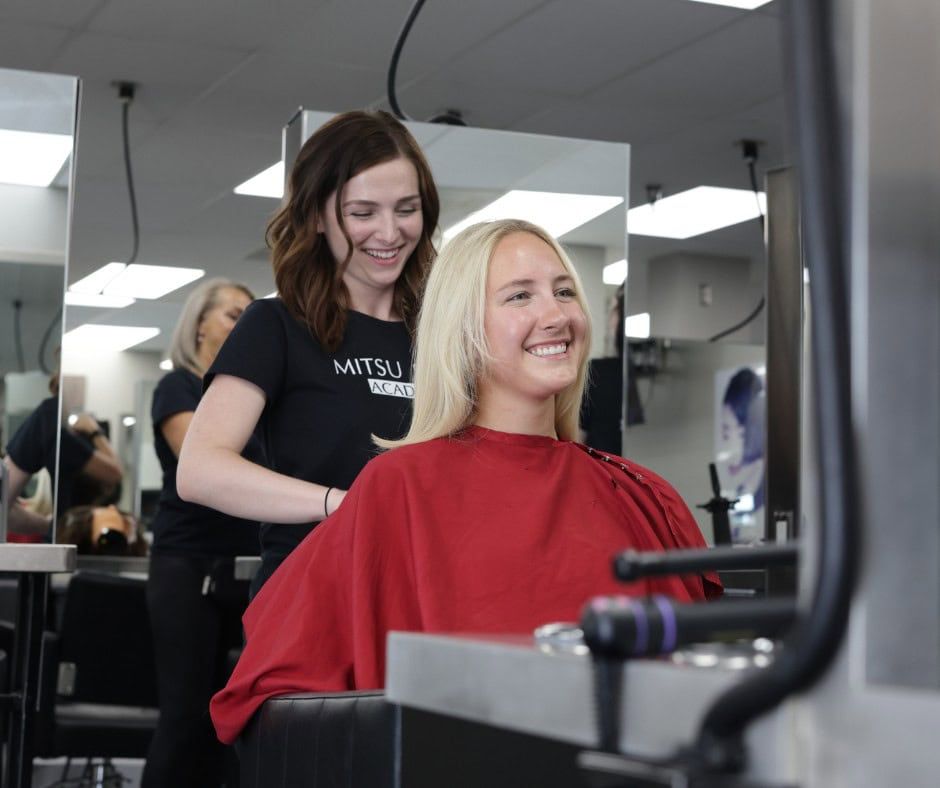 A smiling stylist in a black shirt stands behind a client in a red salon cape at a hair salon.