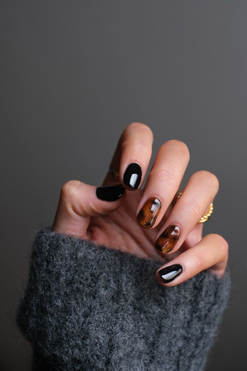 Hand with dark gray sweater sleeve, nails painted black and tortoiseshell, against gray background.