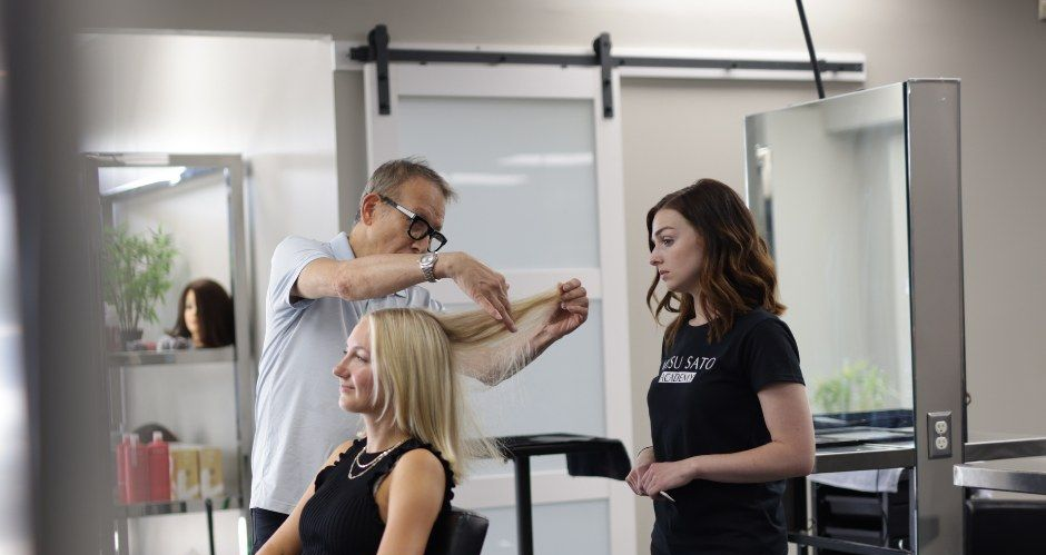 A hairstylist cuts the hair of a seated client while another person observes in a salon.