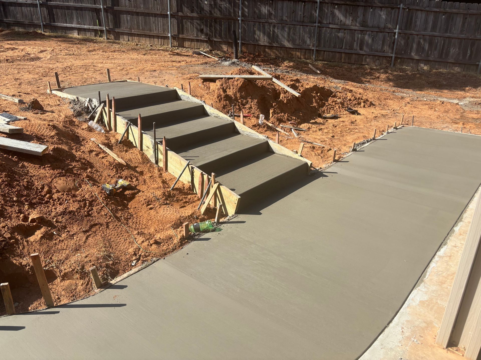 A newly poured concrete patio outside a brick house. Dirt pile in the foreground. Blue sky overhead.