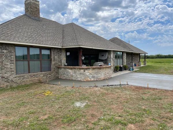 A one-story brick house with a newly poured concrete patio, surrounded by green grass and a cloudy sky.