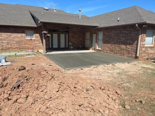 A newly poured concrete patio outside a brick house. Dirt pile in the foreground. Blue sky overhead.