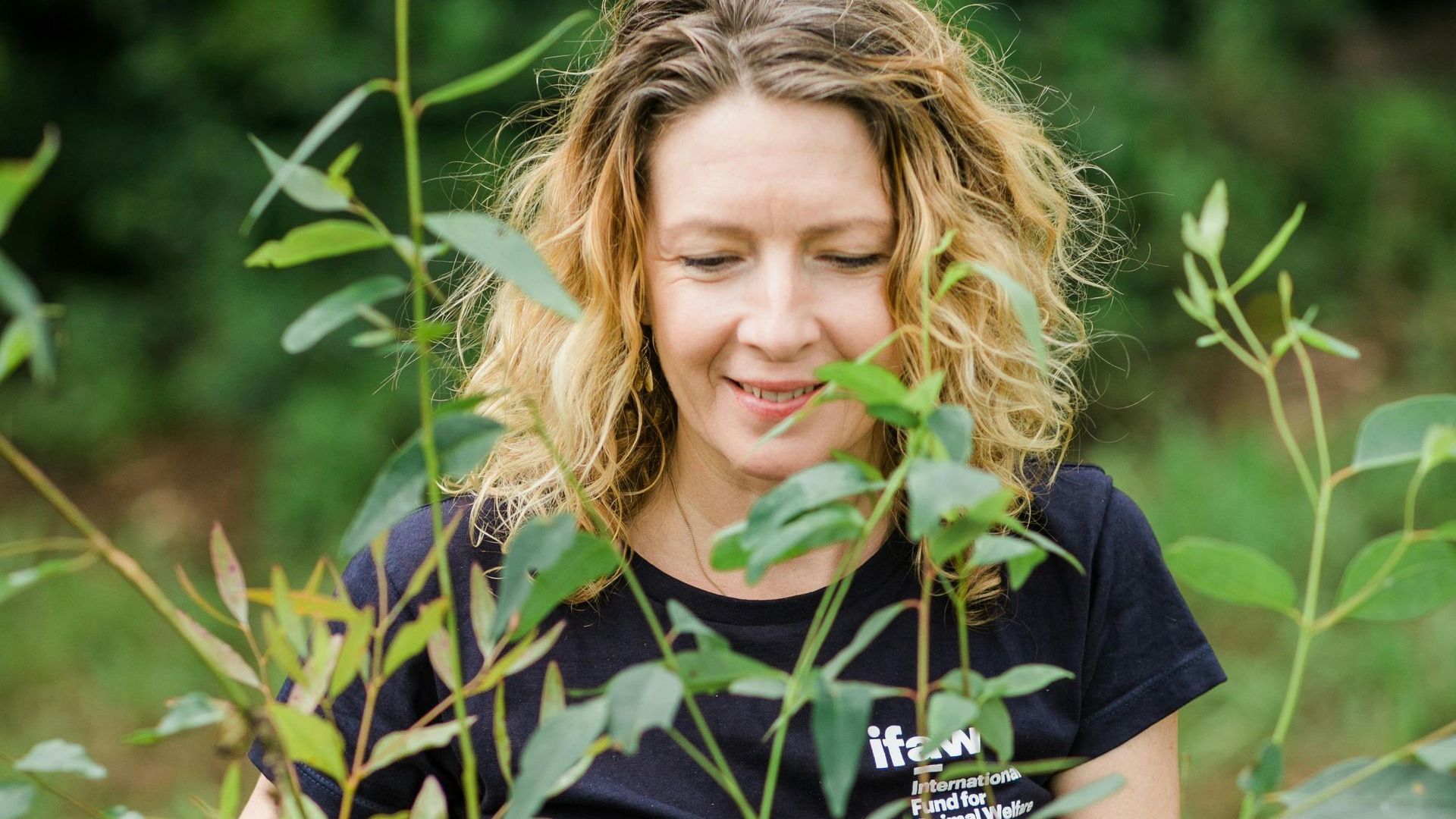 A woman is smelling a plant in the woods.