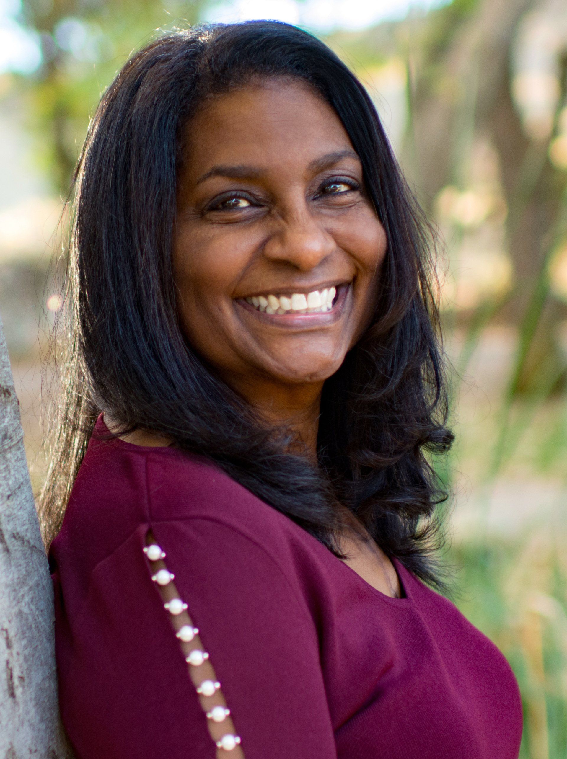 A woman in a red shirt is leaning against a tree and smiling.