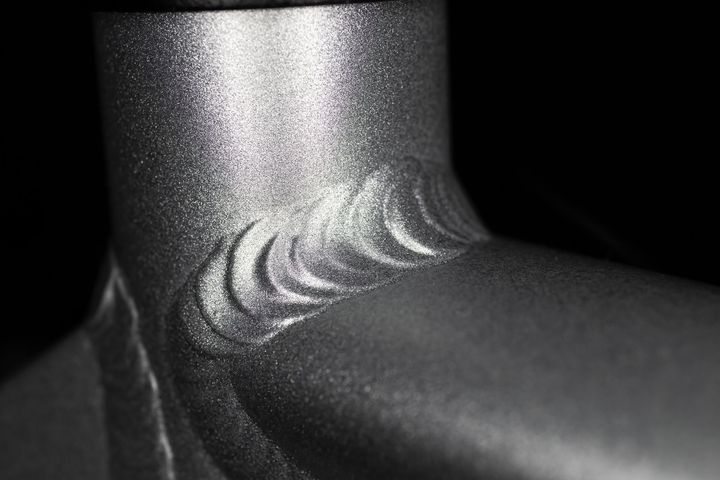 A close-up of a smooth, ripple-pattern aluminum weld connecting two grey metal bicycle frame tubes against a dark background.